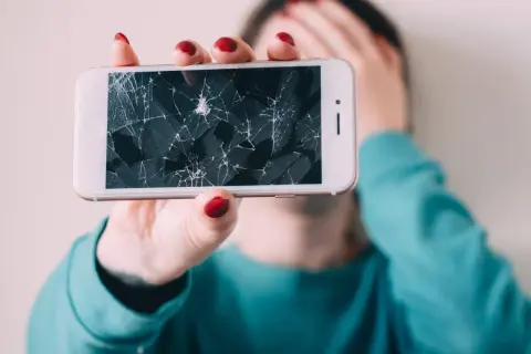 Złamany telefon z pękniętym ekranem, który osoba trzyma przed twarzą, zakrywając ją ręką.