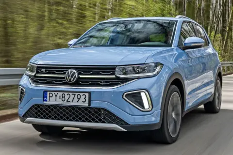 Niebieski VW T-Cross pędzi leśną drogą, odbijając drzewa w szybie.