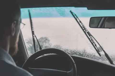 Widok z kabiny samochodu podczas deszczu. Kierowca patrzy na zaparowaną szybę, na której pracują wycieraczki. Zastanawia się, czym czyścić wycieraczki samochodowe, by zapewnić sobie dobrą widoczność.