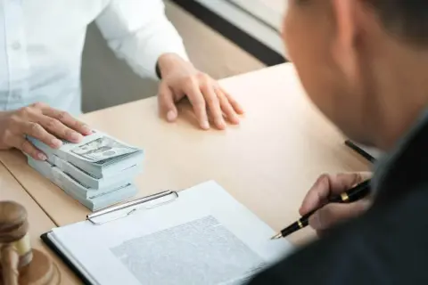 Osoba podpisuje dokumenty, obok stos banknotów. To może być depozyt notarialny.
