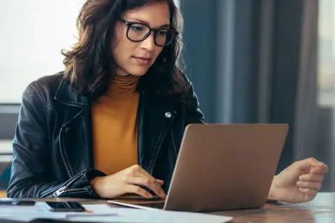 Kobieta w okularach i skórzanej kurtce pracuje na laptopie, szukając informacji, jak sprawdzić lata pracy w ZUS PUE.
