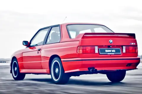 Czerwone BMW E30 Coupe M3 w ruchu, z widocznym spojlerem i podwójnym wydechem.