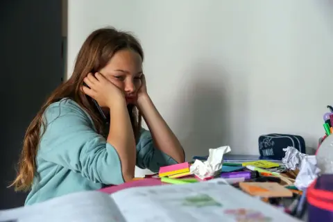 Mädchen sitzt gelangweilt am Schreibtisch, umgeben von Schulmaterialien. Endlich wieder Schule, aber die Motivation fehlt.
