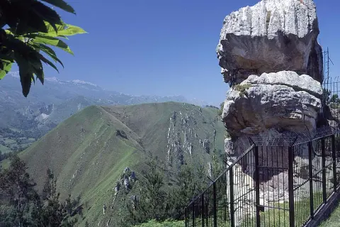 El idolo de peña tu, una roca con forma de perfil humano, vigila un paisaje montañoso verde y sereno bajo un cielo azul.