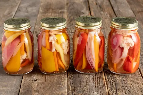 Quattro barattoli di giardiniera fatta in casa, con verdure colorate come peperoni, carote e cavolfiore, su un tavolo di legno rustico.