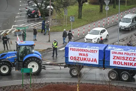 Niebieski traktor ciągnie przyczepę z banerami "Pseudoekolodzy was oszukują" i "Unijna polityka zniszczy rolnika".