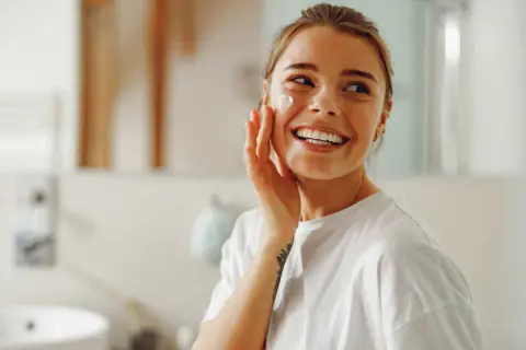 Une femme souriante applique une crème sur sa joue. Elle se prépare pour la journée, peut-être un primer avant ou après crème solaire.