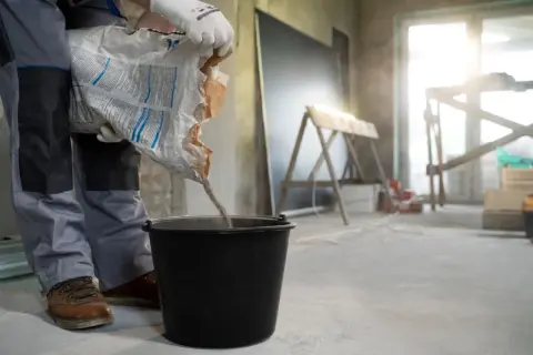 Budowlaniec wsypuje masę do wiadra, przygotowując wylewkę samopoziomującą. Minimalna grubość wylewki samopoziomującej jest kluczowa dla równej podłogi.