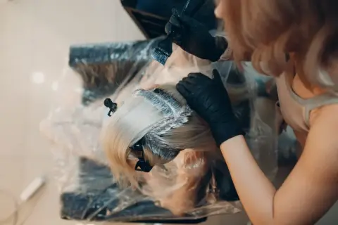 Fryzjerka nakłada farbę na blond włosy. Zastanawiasz się, ile trzymać farbę na włosach? Czas zależy od efektu.