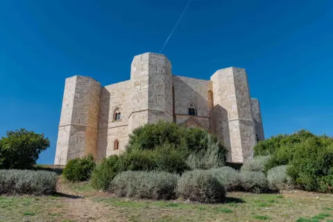 Castel del Monte, un'imponente fortezza ottagonale in pietra chiara, si erge maestoso sotto un cielo azzurro intenso, circondato da vegetazione mediterranea.