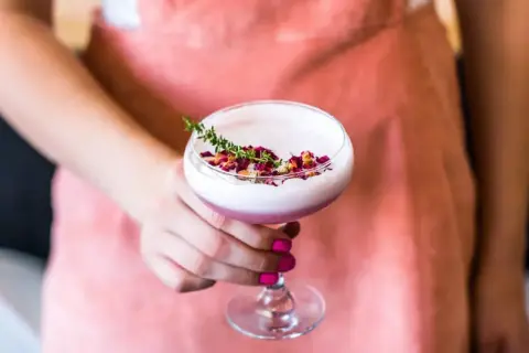 Eleganckie drinki z różowym ginem, ozdobione płatkami róż i gałązką tymianku, prezentowane na tle łososiowego fartucha.
