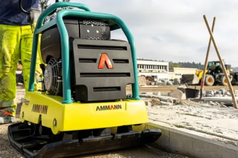 Robotnik na budowie, obok niego stoi żółta zagęszczarka Ammann. Wygląda na to, że zagęszczarka nie jedzie do przodu, bo operator stoi obok.
