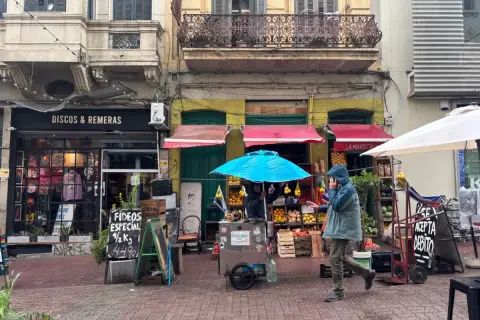 Un hombre con capucha camina por una calle adoquinada en Montevideo, pasando por tiendas de discos y un puesto de frutas.