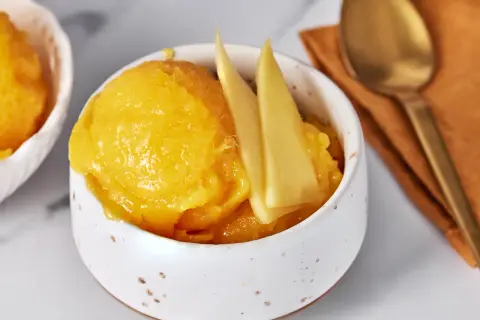 Erfrischendes Mango Sorbet in einer Schale, garniert mit Mangostücken. Daneben ein Löffel auf einem orangen Tuch.