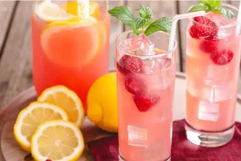 Två glas med rosa lemonad, hallon, is och mynta. Citroner och en karaff i bakgrunden ger härliga sommar vibes.