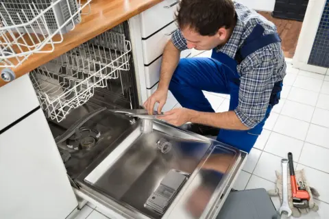 Instalator w niebieskim kombinezonie pokazuje, jak podpiąć zmywarkę, pracując przy jej wnętrzu. Narzędzia leżą obok.