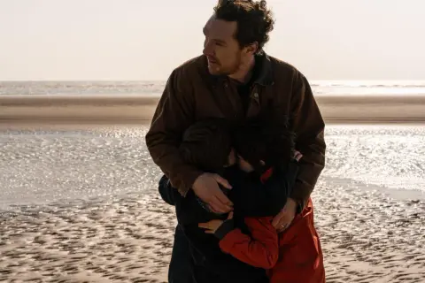 En man kramar sina barn på stranden. En scen som känns som en av de där comfort movies man vill se om och om igen.