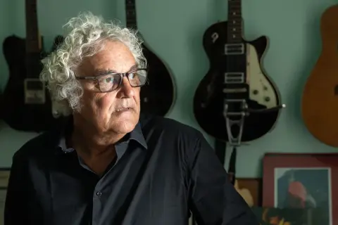 Massimo Bubola, con i suoi capelli ricci e bianchi, guarda pensieroso tra le chitarre appese al muro, testimoni silenziose delle sue canzoni.