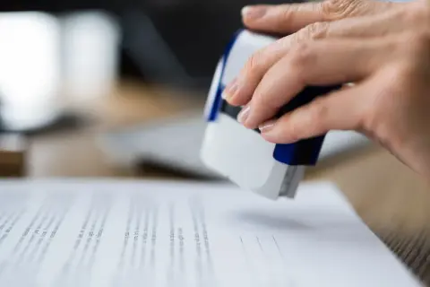 Dłoń z pieczątką nad dokumentami, które mogą być pozwolenie na budowę.