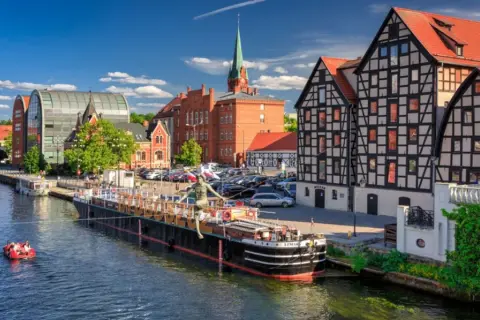 Nad rzeką w Bydgoszczy, jednym z 20 największych miast w Polsce, cumuje barka. W tle zabytkowe kamienice i nowoczesna architektura.