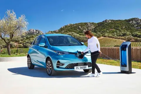 Une femme branche sa Renault Zoé bleue à une borne de recharge. Le temps de recharge zoe 22kw est rapide, parfait pour profiter du paysage.