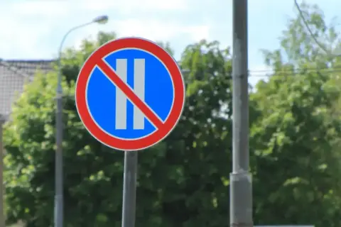 Znak drogowy: zakaz postoju. Dwa pionowe białe paski na niebieskim tle, przekreślone czerwoną linią.