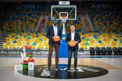 Dos hombres posan con balones de baloncesto en una cancha vacía. Detrás de ellos, una pila de cajas de frutas y asientos de colores. Un guiño al club baloncesto Gran Canaria.