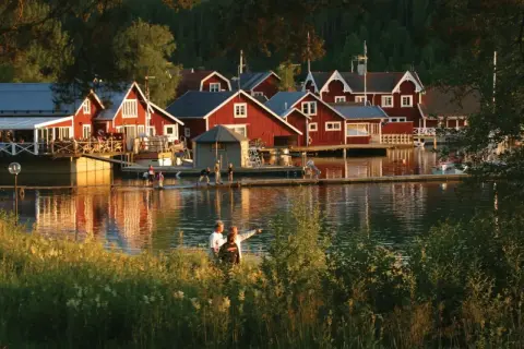 Solnedgång över Norrfjällsviken med röda sjöbodar och folk som njuter av kvällen vid vattnet.
