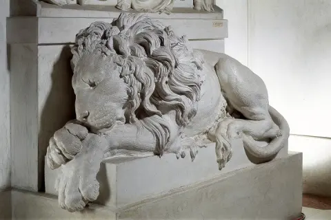 Leone scolpito, simbolo di forza, riposa ai piedi della tomba di Clemente XIII. La sua criniera fluente e le zampe possenti trasmettono un senso di maestà e quiete eterna.