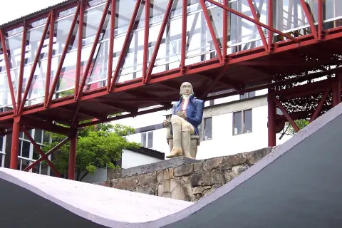 Estatua de un hombre sentado frente a la fábrica de Sargadelos, bajo un puente rojo y acristalado.