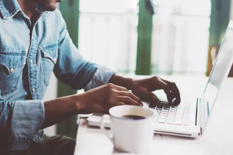 Mężczyzna w dżinsowej koszuli pisze na laptopie, obok stoi filiżanka kawy.