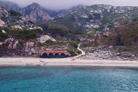 Spiaggia selvaggia con rocce e vegetazione, un edificio storico e montagne nebbiose sullo sfondo. Qui si trova l'isola di Montecristo.