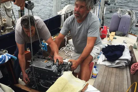 Due uomini lavorano alla manutenzione del motore di una barca a vela, sollevandolo con una catena.
