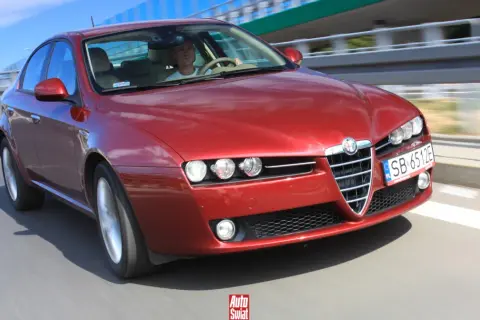 Czerwone Alfa Romeo 159 1.9 JTD pędzi po drodze. Kierowca w białej koszulce pewnie trzyma kierownicę.