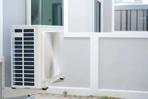 Unité extérieure d'une pompe à chaleur appartement, fixée sur un mur blanc. L'appareil est blanc avec des grilles bleues et un ventilateur visible.