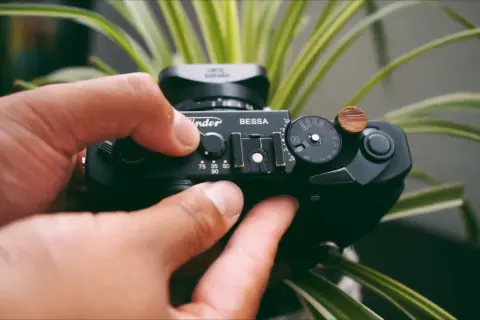 Hands adjust the settings on a black Bessa R2A camera, surrounded by lush green foliage.