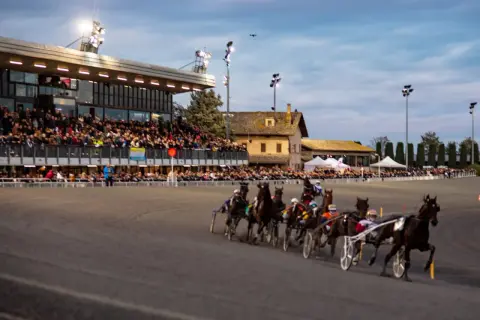 Corsa di cavalli al trotto all'ippodromo Montegiorgio, con pubblico sugli spalti e cavalli in azione sulla pista.