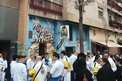 Gente reunida en la calle, algunos con chaquetas blancas y amarillas, celebrando la cincomarzada frente a un edificio con grafitis.