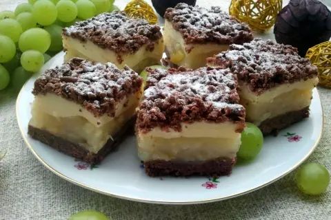 Szybkie ciasto z gruszkami i budyniem, posypane cukrem pudrem, na talerzu z winogronami.
