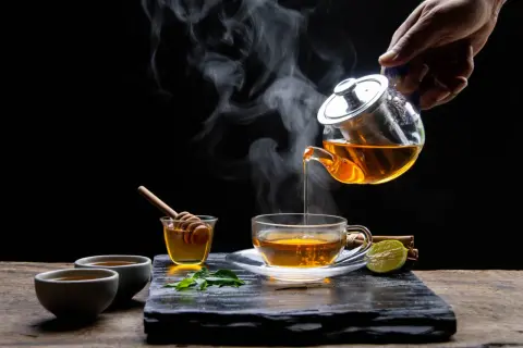 Une main verse un thé rose fumant dans une tasse. Du miel, de la menthe et de la citron vert accompagnent cette boisson réconfortante.