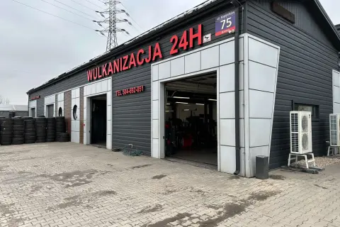 Budynek wulkanizacji z napisem "Wulkanizacja 24H", stosy opon i otwarte bramy garażowe.