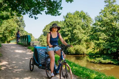 Mujer pedalea por las vías verdes de Francia con un remolque para niños, disfrutando del paisaje junto al canal.