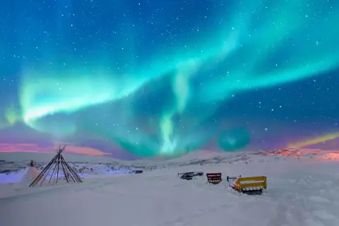 Niesamowita zorza polarna nad śnieżnym krajobrazem Norwegii. W oddali widać tipi i sanie, gotowe na nocną przygodę.