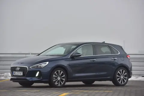 Ciemnoniebieski Hyundai i30 na parkingu. Kierowca w okularach siedzi za kierownicą, gotowy do wymiany żarówki.