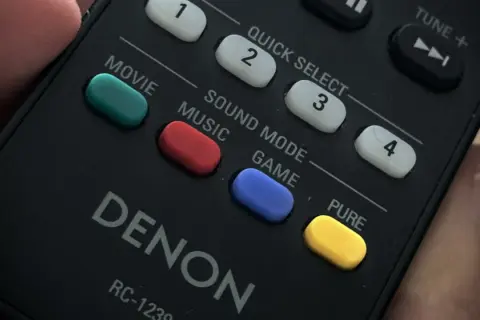 Dłoń trzyma pilot Denon. Widoczne przyciski: Movie, Music, Game, a także żółty przycisk PURE.