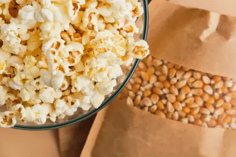 Miseczka pełna puszystego popcornu obok ziaren kukurydzy w papierowej torbie. Czy popcorn jest lekkostrawny? Zależy od dodatków!