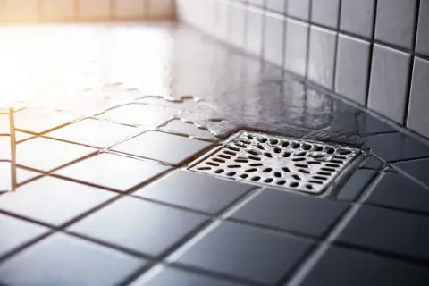 Acqua stagnante vicino allo scarico doccia puzza. Le piastrelle scure e bagnate creano un'atmosfera poco invitante.