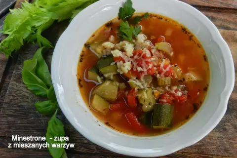 Miseczka zupy minestrone z warzywami i startym serem, obok łodygi selera i liście bazylii.