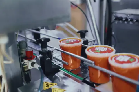 Linia produkcyjna z zupami w plastikowych kubkach z czerwonymi etykietami.