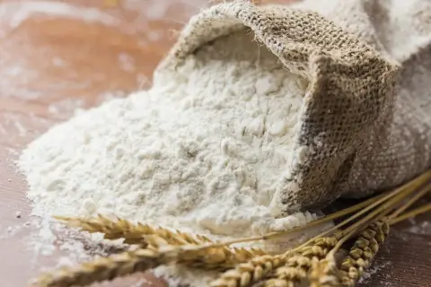 Wsypana z worka mąka pszenna i kłosy zbóż. To właśnie z nich powstaje gluten, kluczowy składnik wypieków.